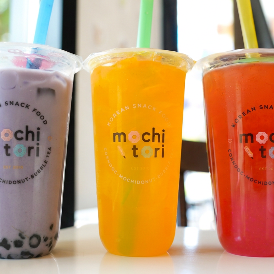 Three Mochi Tori bubble tea cups in purple, orange, and red flavors sit on a café counter.